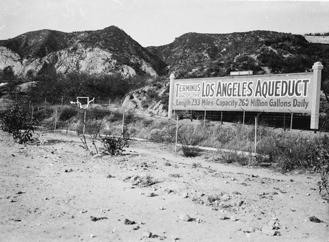 (n.d.)^# - Terminus of the Los Angeles Aqueduct - This photograph shows a sign advertising the terminus of the Los Angeles Aqueduct, which had been completed in November, 1913. The sign was located near where present day Interstate 5 runs just east of the Van Norman Reservoir in Sylmar. In the background to the North are the San Gabriel Mountains, just east of the Newhall Pass. To the left of the sign can be seen the original water cascade that marked the terminus of the Aqueduct. The sign states "Terminus of the Los Angeles Aqueduct. Length 233 Miles. Capacity 260 Million Gallons Daily".